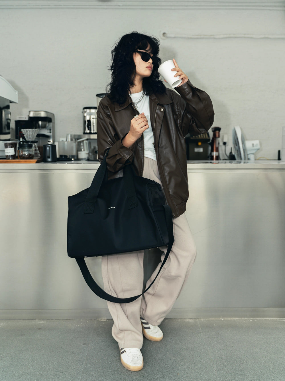 woman-leaning-on-stainless-steel-coffee-bar-holding-coffee-in-left-hand-elbow-carrying-black-kamamuta-pet-shadow-stealth-whisper-square-bag-carrier