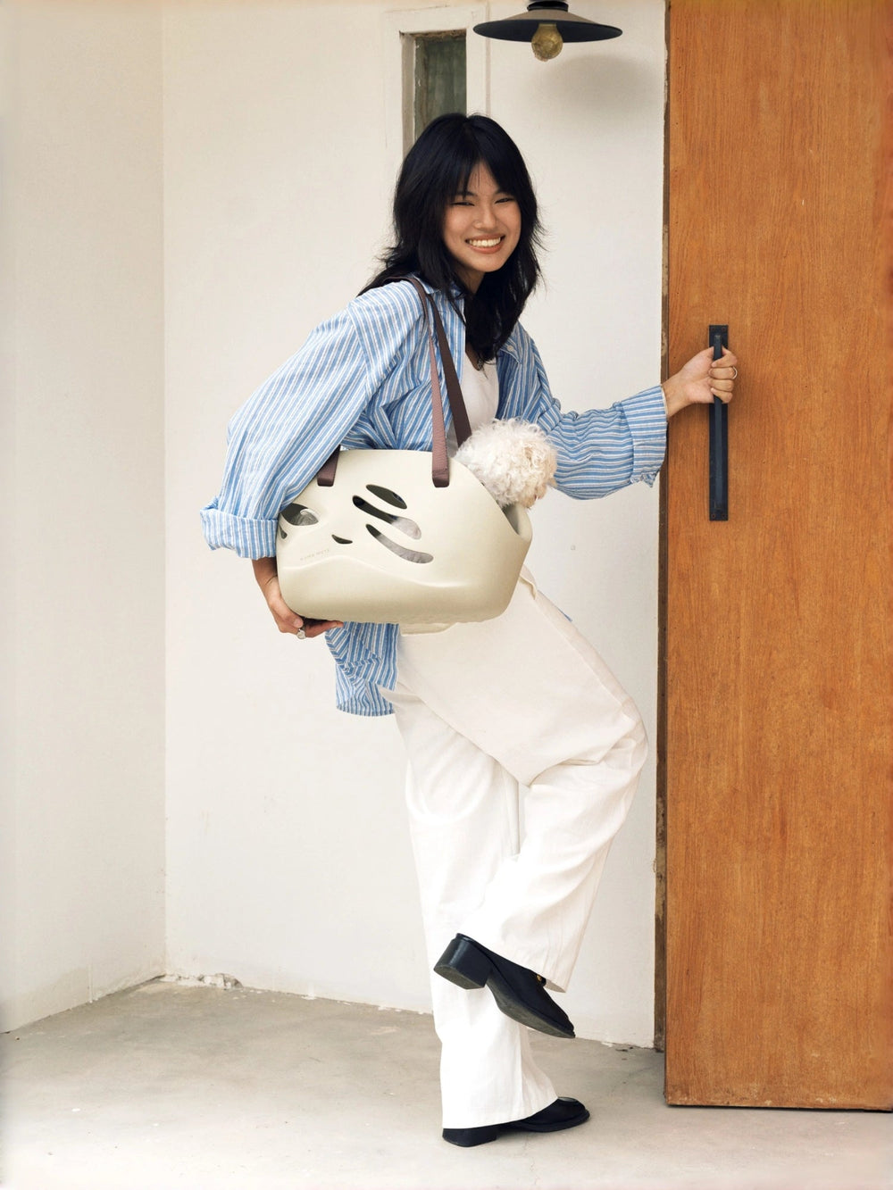 fashionable-young-woman-holding-door-handle-looking-back-at-camera-with-kamamuta-wave-pet-tote-bag-on-right-shoulder-carrying-white-maltese-dog